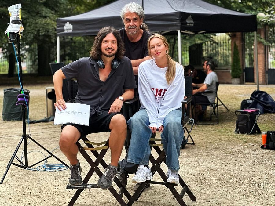 En una escena de rodaje al aire libre, tres personas, vestidas con ropa informal, descansan en sillas de director. Un guion con la anotación 