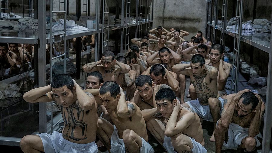 Numerosos presos, agachados y con las manos en la cabeza, en espacio confinado con literas metálicas. Visten ropa interior blanca; muchos muestran tatuajes.