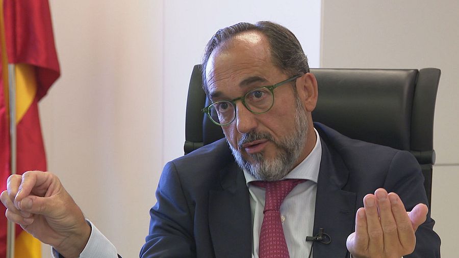 Fiscal de Sala Coordinador de Seguridad Vial sentado, gesticulando, con traje oscuro, camisa blanca y corbata granate. Expresión seria y concentrada. Bandera de España visible.