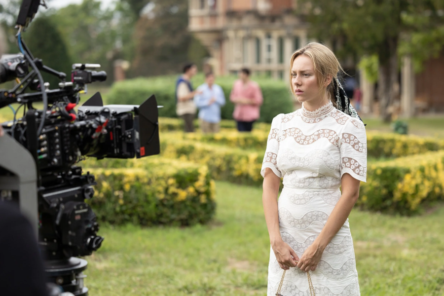 Actriz con vestido blanco y bolso pequeño, en rodaje cinematográfico.  Ambiente formal, cámara profesional y equipo de filmación visibles.