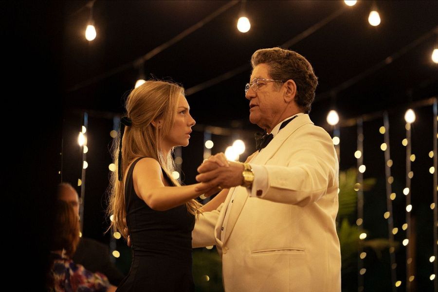 Escena de baile entre dos actores; ella con vestido negro y él con traje blanco, en un ambiente nocturno y festivo.