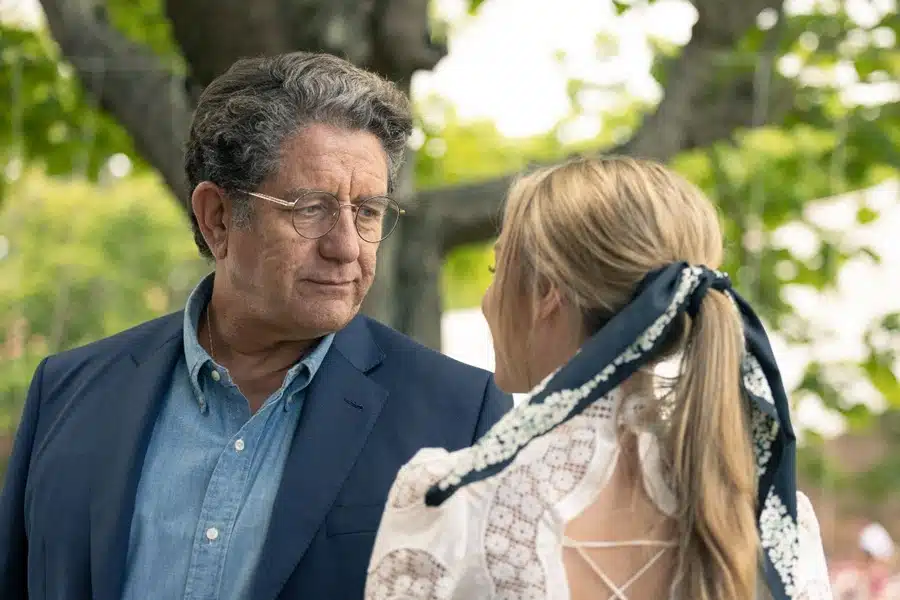 Conversación seria al aire libre entre dos personas; él, en traje azul marino, ella, con blusa blanca y pañuelo azul.  Entorno natural.