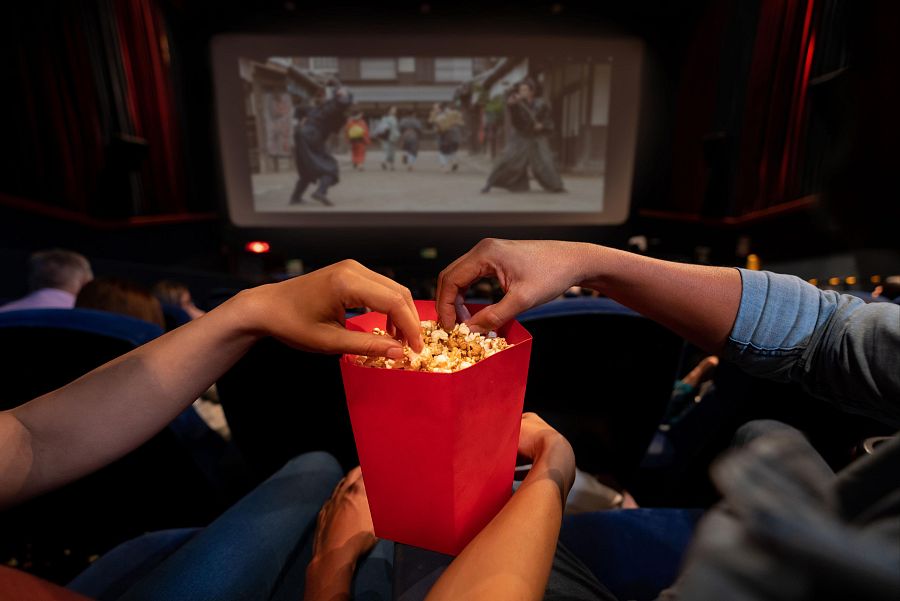 Dos personas toman palomitas de un cubo rojo en una sala de cine mientras se proyecta una película ambientada en el Japón feudal, con guerreros.