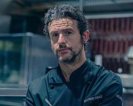 Chef con uniforme negro y barba canosa, sosteniendo un utensilio. Expresión seria, mirando a cámara.  Imagen promocional de comida sin gluten.