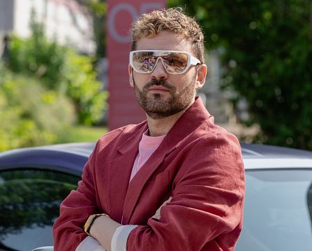 Hombre con barba y gafas de sol, americana roja y camiseta rosa, posa con los brazos cruzados. Anuncio comercial sin gluten.