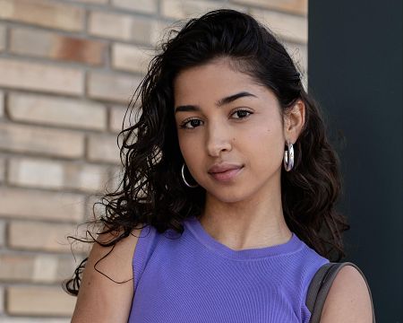 Joven con cabello oscuro y rizado, expresión seria, camiseta morada y grandes aros plateados. Fondo con pared de ladrillo beige y columna gris.