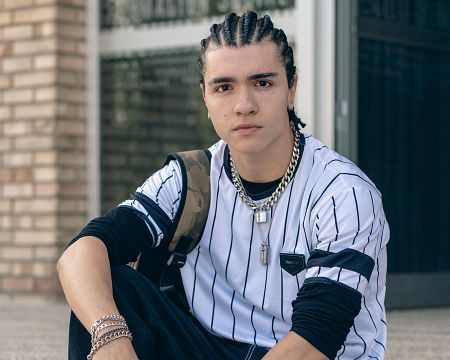 Joven con trenzas, camiseta blanca con rayas azules y negras, mochila camuflaje y varias cadenas y pulseras plateadas, sentado con expresión seria mirando a cámara.