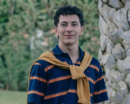 Joven con polo a rayas y jersey mostaza en entorno natural, imagen promocional de producto 'COM sin gluten'.