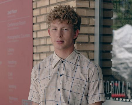 Joven de cabello rubio rizado, camisa a cuadros, expresión neutra, mirando a cámara. Fondo con pared de ladrillo y cartel rosa con directorio.