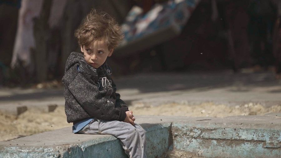 Niño sentado en bordillo bajo, expresión seria, sudadera gris con capucha y pantalones grises. Fondo desenfocado, ambiente precario.