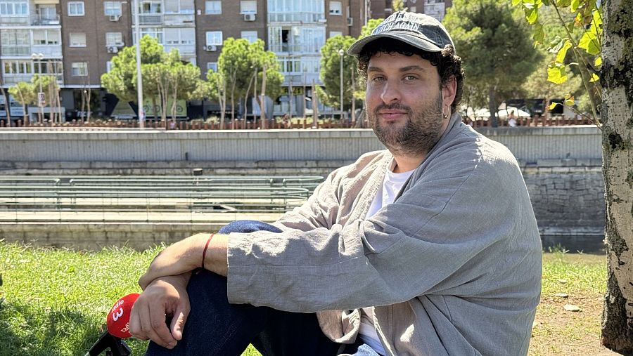 Músico sentado al aire libre, con gorra y camisa gris, sostiene micrófono con logo de Radio 3. Entrevista en entorno natural, cerca de un río.