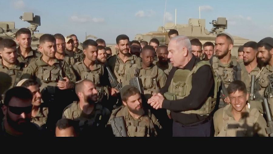 Netanyahu, con chaleco antibalas, estrecha la mano de un soldado israelí, rodeado de otros uniformados en una zona que parece ser militar.  Se aprecia una atmósfera de camaradería.