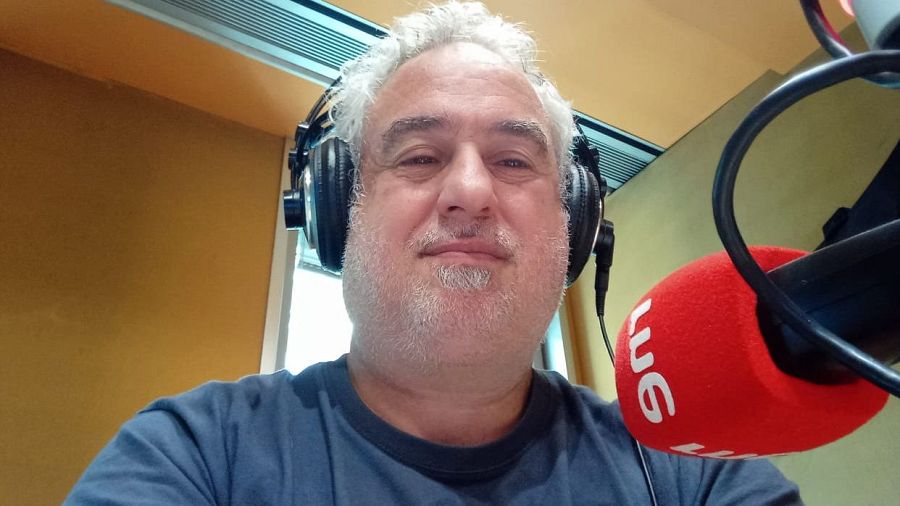 Periodista con auriculares y micrófono en estudio de radio.  Pelo y barba canosos.  Fondo mostaza, logo RNE visible.