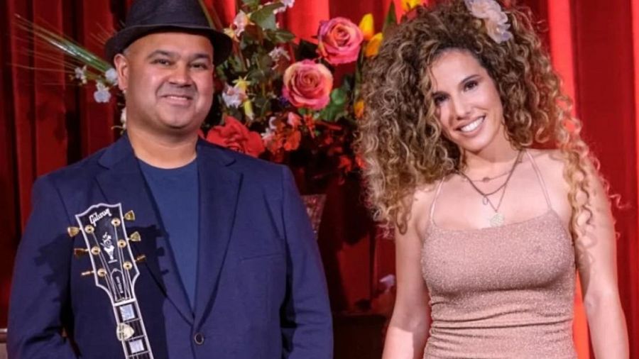 Dos personas, una con sombrero y guitarra, la otra con vestido brillante, posan ante un fondo rojo con flores.