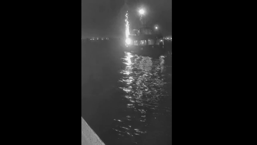 Barco iluminado en el agua, fotografiado de noche con un dron.  Luces brillantes reflejadas en el agua oscura. Perspectiva aérea o desde otro barco.