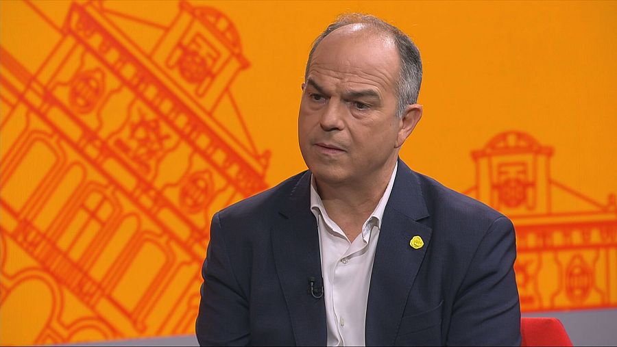 Entrevista a Jordi Turull a 'Aquí Parlem' de TVE Catalunya. Vesteix amb vestit fosc i camisa blanca, amb un fons taronja i silueta d'edifici.