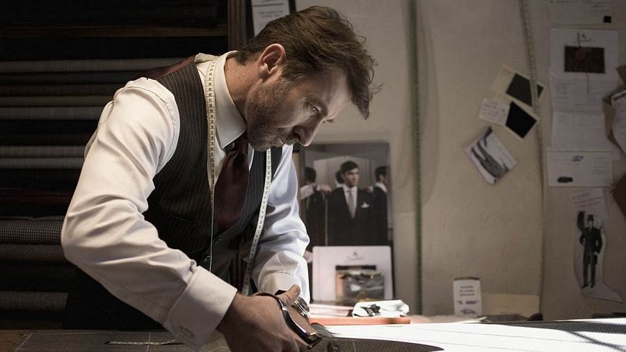 Sastre cortando tela en su taller.  Vestimenta: camisa blanca, chaleco oscuro y corbata granate.  Entorno con rollos de tela y fotografías en la pared.