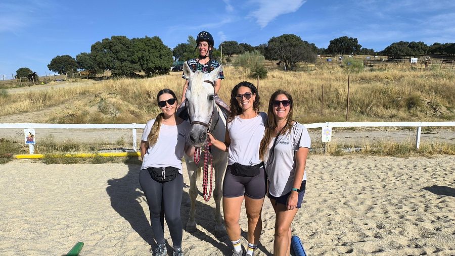 Jinete a caballo blanco en pista de arena con tres mujeres cerca.  Entorno rural.  Imagen relacionada con actividades ecuestres.