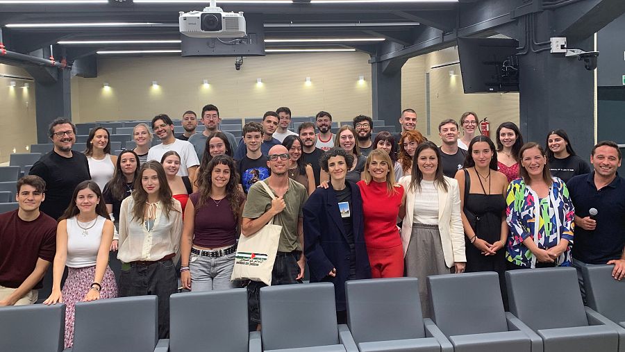 Grupo de personas, entre 20 y 30 años, posando en un aula.  Un hombre sostiene un micrófono, una mujer viste un traje rojo.  Se ven bolsas con logo.