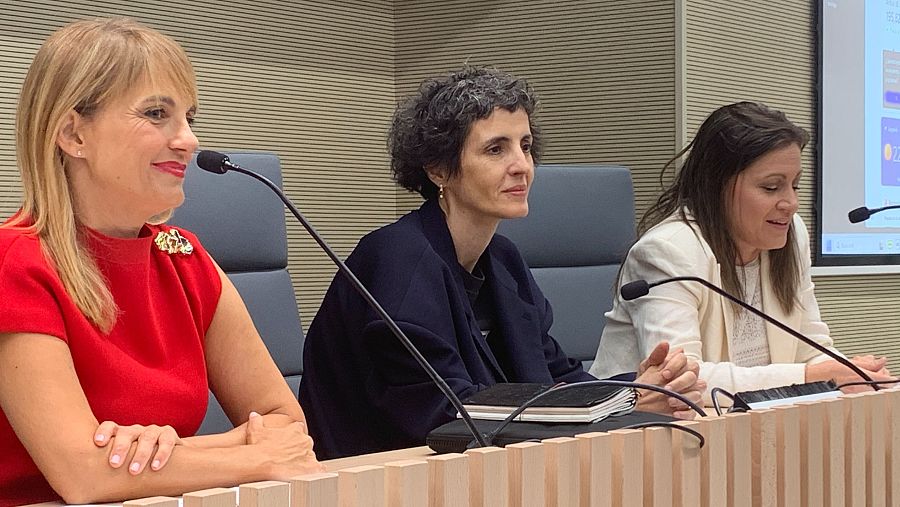 Conferencia de prensa: tres mujeres con micrófonos individuales sentadas tras una mesa de madera clara.  Una lleva blusa roja, otra chaqueta oscura y la tercera una clara.