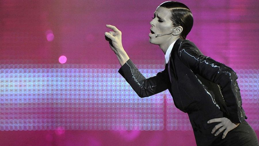 Imagen de perfil de Bimba Bosé actuando: traje negro, camisa blanca, micrófono, pelo recogido y fondo rosa con luces circulares.