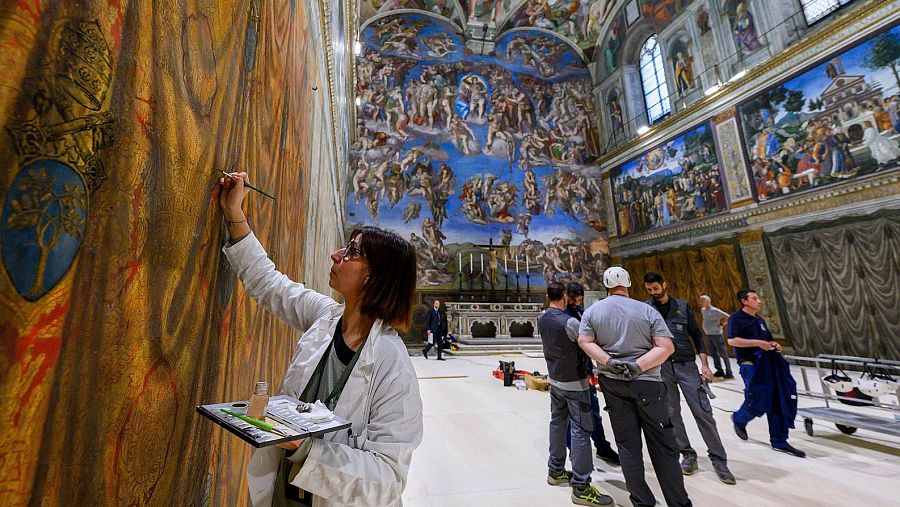 Restauración de un fresco en la Capilla Sixtina: una mujer con bata blanca trabaja con pinceles y paleta, rodeada de andamios y otros restauradores.