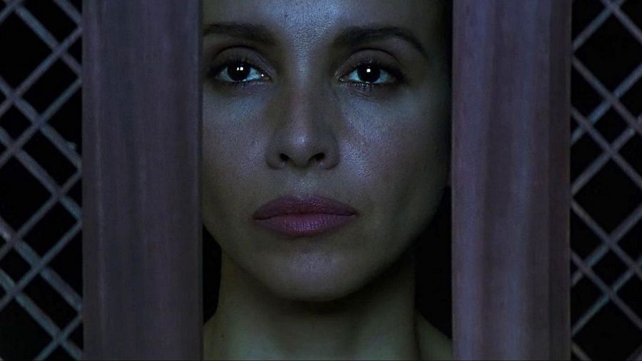 Retrato de Ana Belén con expresión seria, parcialmente oculto tras una celosía de madera y malla metálica al fondo. Iluminación tenue.