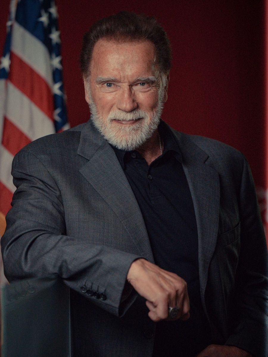 Imagen cercana de Arnold Schwarzenegger sonriendo, con americana gris sobre camisa oscura, cabello y barba canosos. Fondo con bandera estadounidense.
