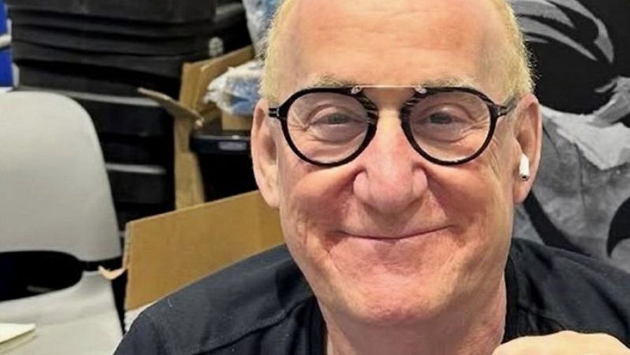 Primer plano de Jeph Loeb, escritor de cómics, con gafas y auricular inalámbrico; fondo con sillas y cajas, indicando un evento.