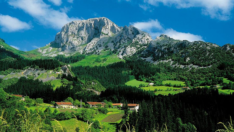 Vista del Monte Mugarra en el Parque Natural de Urkiola, con prados verdes, bosques de pinos y caseríos vascos en primer plano.