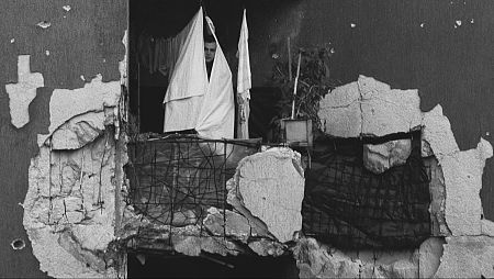 Hombre escondido tras sábana en balcón de edificio en ruinas.  Su rostro muestra miedo;  a pesar de la destrucción, hay una planta y ropa tendida.