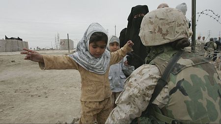 Niña con ropa clara y pañuelo levanta brazos ante soldado con casco.  Fondo árido con alambrada y otras personas.  Posible control militar.