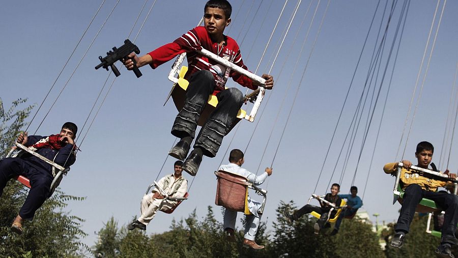 Niños en un columpio de un parque de atracciones; uno porta una metralleta de juguete.  Contrasta la diversión infantil con la seriedad de un arma, aunque sea de juguete.