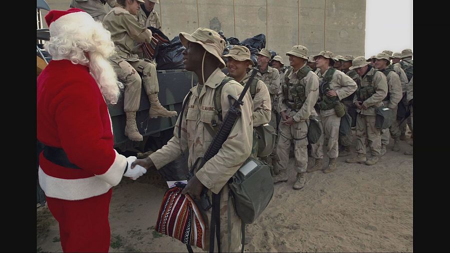 Papá Noel entrega un regalo a un soldado afroamericano en uniforme, rodeado de otros soldados en un ambiente desértico.  Escena navideña en un contexto militar.
