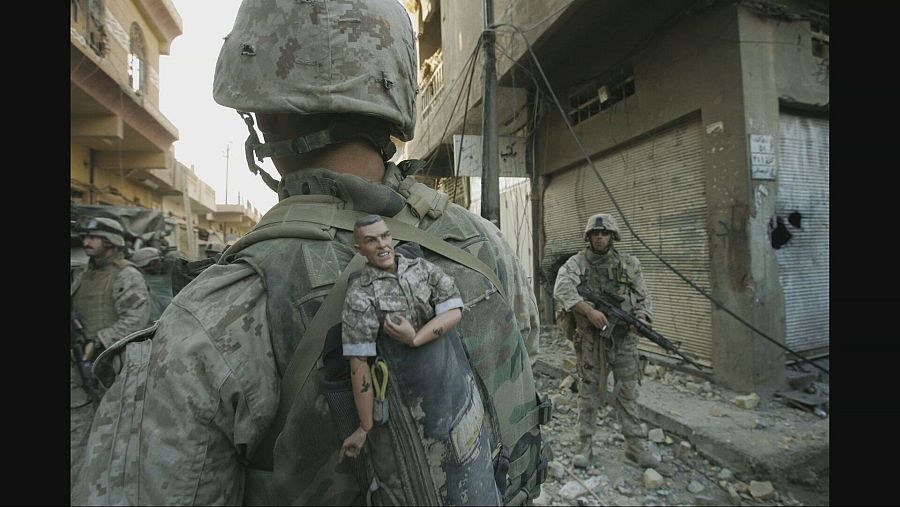 Marine con muñeco en la mochila en zona de combate urbana.  Edificios dañados y otros soldados al fondo.  Imagen en blanco y negro.