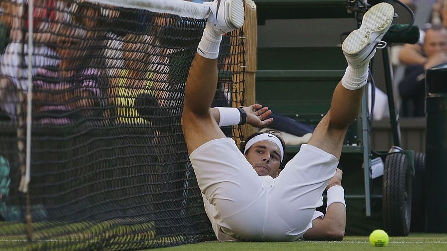 Tenista en pista de tenis, vestido de blanco, realiza estiramientos con las piernas sobre la red y una pelota a sus pies. Fondo con público y red.