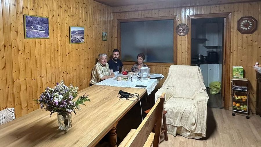 Tres personas comparten una comida en una casa de madera, con una atmósfera cálida y hogareña.  La mesa está decorada con flores y hay comida, platos y vasos.