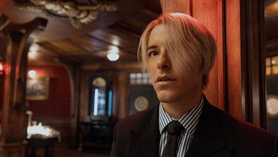 Imagen de Taz Skylar con traje oscuro y camisa a rayas, expresión seria, mirando a la izquierda. Fondo con decoración clásica: madera oscura, pared roja, mesa con mantel blanco.