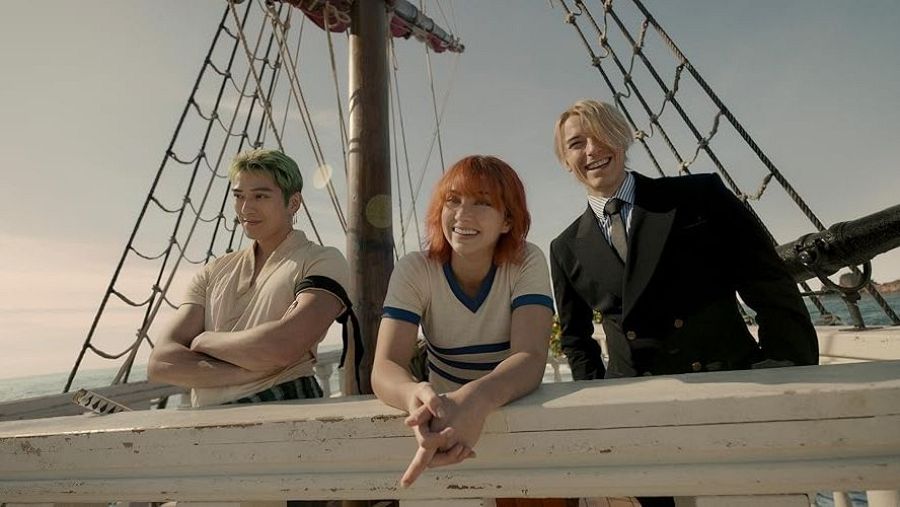 Tres actores, vestidos de época, sonríen en un barco de madera, mirando hacia el mar.  Escena de una serie de televisión.