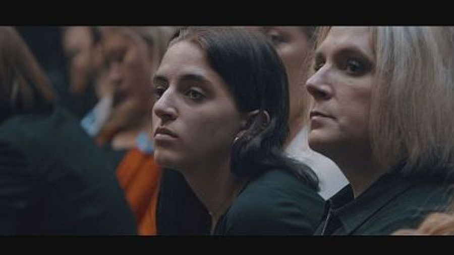 Dos mujeres en una audiencia observan algo fuera de la imagen. Una tiene cabello castaño y pendiente dorado; la otra, cabello rubio con canas y camisa oscura.  Ambiente serio y atento.