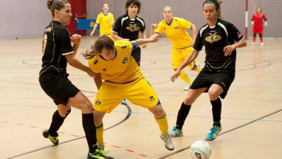 Imagen de un partido de fútbol sala femenino en una cancha interior. Dos jugadoras, una con uniforme negro y otra con uniforme amarillo, disputan el balón. En el fondo, se observan más jugadoras participando en el juego.