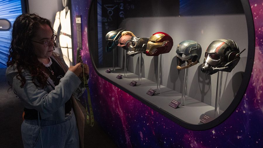Una mujer con gafas y cabello rizado examina una vitrina que contiene cinco cascos de superhéroes, posiblemente de los Vengadores. La mujer, vestida de forma casual, sostiene un teléfono móvil y parece estar fotografiando los cascos.