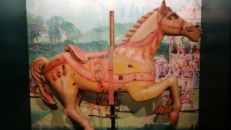 Un caballo de tiovivo de color crema, con detalles en rosa y rojo, se muestra en una pose de galope. La silla de montar es rosa y la pintura desgastada sugiere que es un objeto antiguo, con una barra vertical que lo atraviesa.