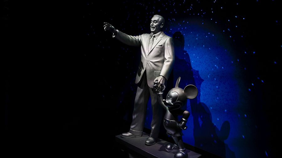 Una estatua de Walt Disney, vestido con traje, y Mickey Mouse, posan juntos. El fondo oscuro con estrellas crea un ambiente mágico.