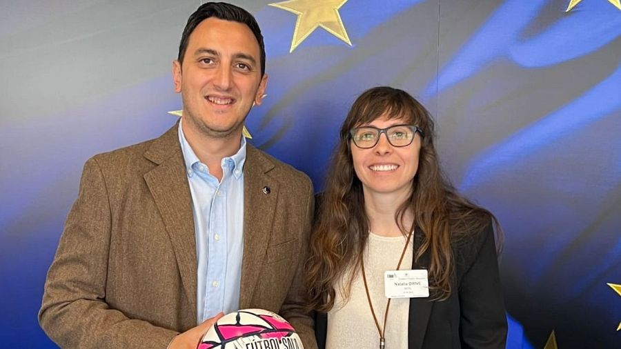 Dos personas se encuentran frente a la bandera de la Unión Europea. Un hombre con un balón de fútbol sala y una mujer con una identificación con nombre posan para la foto.