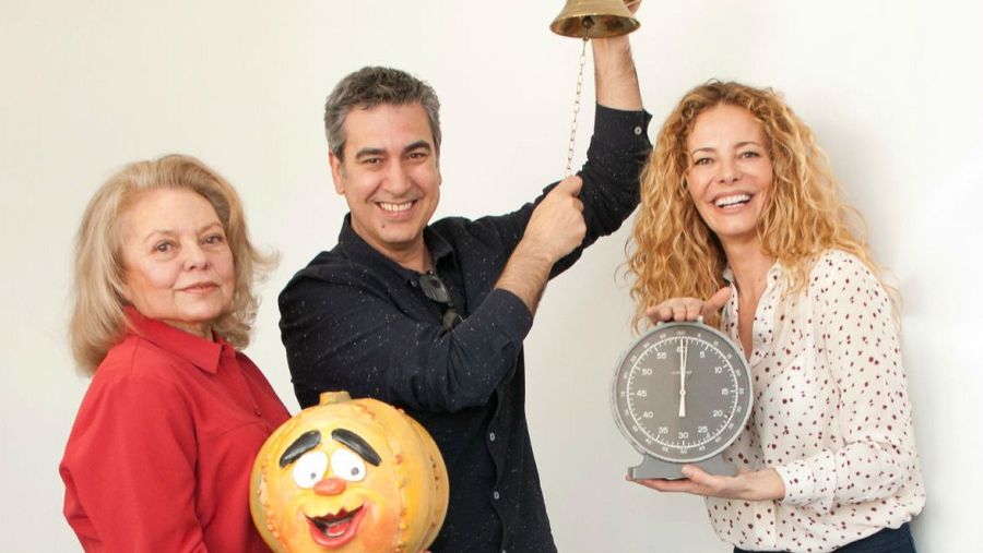 Tres presentadores sonríen frente a un fondo blanco. Una mujer con jersey rojo posa junto a una calabaza sonriente, mientras que otra sujeta un reloj de gran tamaño. Un hombre levanta una campana dorada.