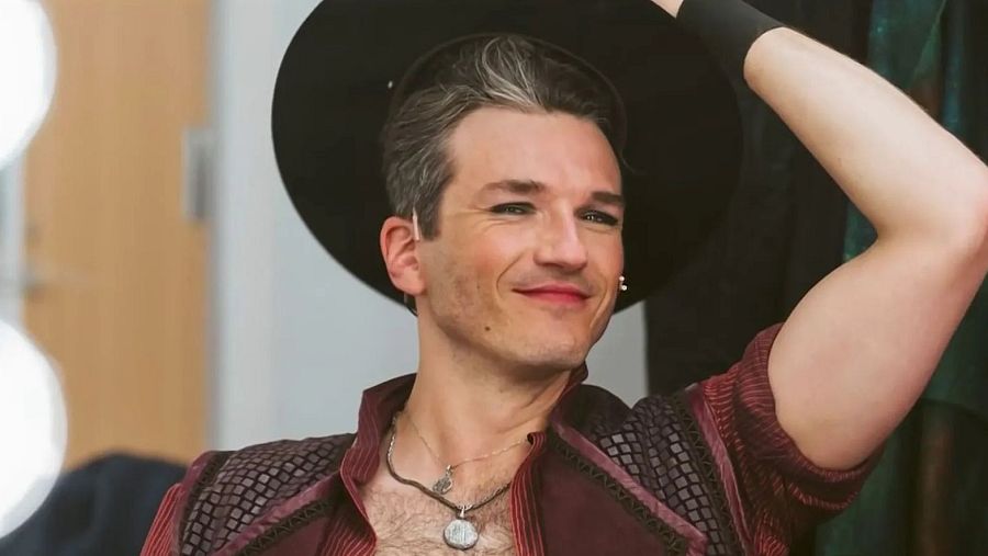 Pepe Muñoz, con maquillaje marcado y sombrero negro, posa con un vestuario que combina burdeos y negro, adornado con collares.