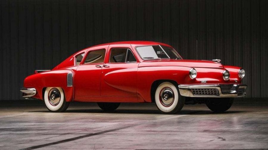 Un coche clásico rojo, un Tucker 48, se exhibe en un estudio. El vehículo, con neumáticos de banda blanca, faros redondos y parrilla cromada, destaca por su diseño aerodinámico y elegante.