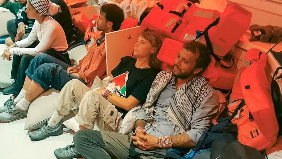 Un grupo de activistas, entre ellos una persona con cabello rubio corto y otra con barba y pañuelo, se encuentran sentados en el suelo de una embarcación, rodeados de chalecos salvavidas.