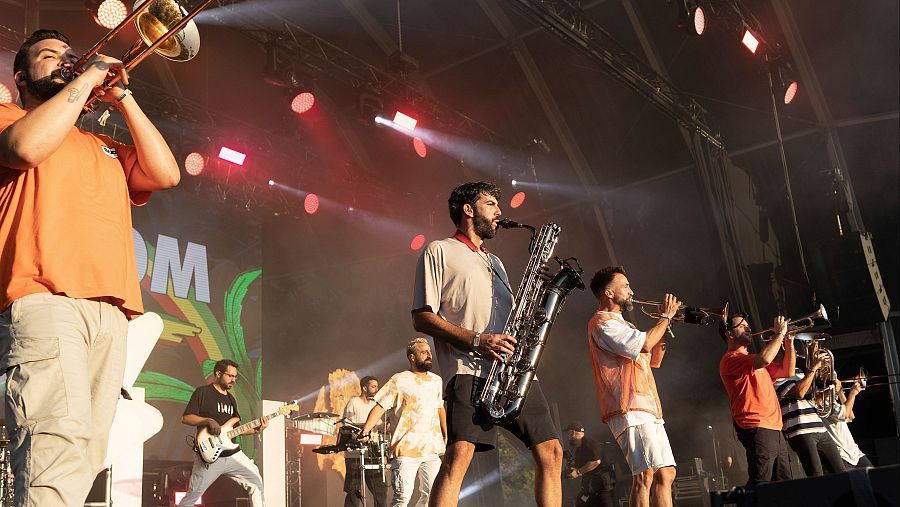 En un escenario iluminado, la banda La Fúmiga actúa durante la clausura de un festival en 2023. Se observa a un saxofonista en primer plano, acompañado por otros músicos tocando diversos instrumentos.